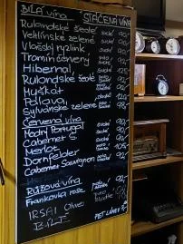 Křídová tabule s ručně psaným seznamem stáčených vín a cenami, nad ní klobouk a staré hodiny